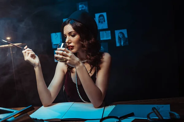 Attractive woman in fascinator hat holding mouthpiece and glass of cognac in dark office — Stock Photo