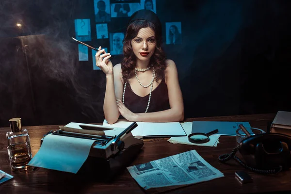 Pretty woman in dress with mouthpiece sitting at table with typewriter in dark office — Stock Photo