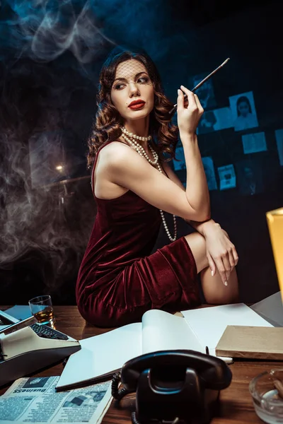 Beautiful young woman in dress holding mouthpiece while sitting on table in dark office — Stock Photo