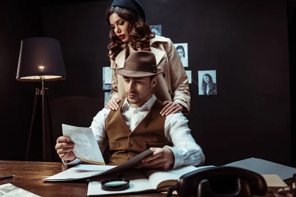 Detective and beautiful woman in trench coat looking at photo in dark office — Stock Photo