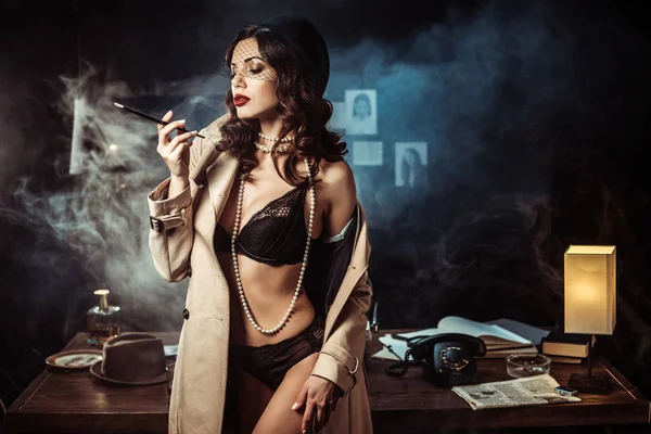 Сексуальна дівчина в чорній білизні і траншеї пальто тримає мундштук в темному офісі — стокове фото