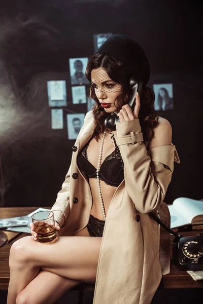 Mulher sexy em lingerie preta e casaco de trincheira segurando vidro de conhaque enquanto conversa no telefone — Fotografia de Stock