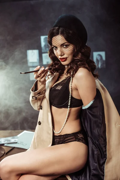Femme sexy en sous-vêtements et trench coat tenant embout buccal tout en étant assis sur la table au bureau — Photo de stock