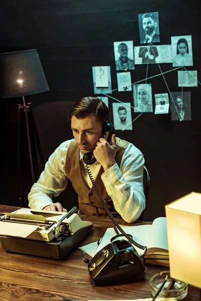 Detective sitting at table with typewriter and talking on telephone in office — Stock Photo