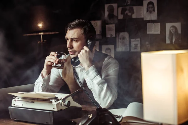 Detective talking on telephone and drinking cognac in office — Stock Photo