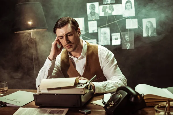 Pensive detective in white shirt sitting at table with typewriter in dark office — Stock Photo