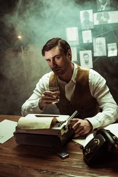 Détective tenant un verre de cognac tout en utilisant une machine à écrire dans un bureau sombre — Photo de stock