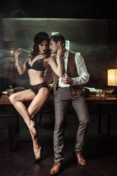 Sexy woman in black lingerie sitting on wooden table and flirting with detective in office — Stock Photo