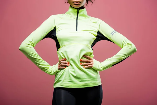 Vista recortada de la deportista afroamericana en chaqueta de atletismo posando con las manos akimbo aislado en rosa - foto de stock