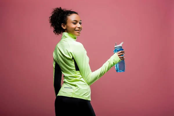 Beautiful smiling african american sportswoman in track jacket with sport bottle looking at camera isolated on brown — Stock Photo