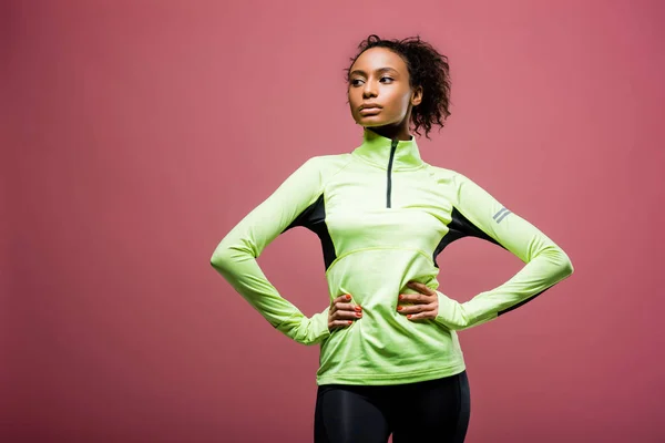 Hermosa afroamericana deportista en chaqueta de atletismo con manos akimbo posando aislado en rosa - foto de stock