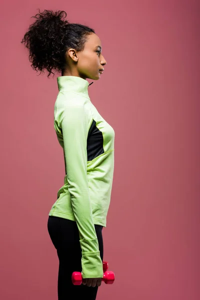 Vista lateral de la hermosa deportista afroamericana sosteniendo mancuerna aislada en marrón - foto de stock