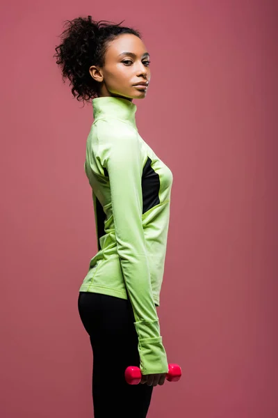 Beautiful african american sportswoman holding dumbbell and looking at camera isolated on brown — Stock Photo