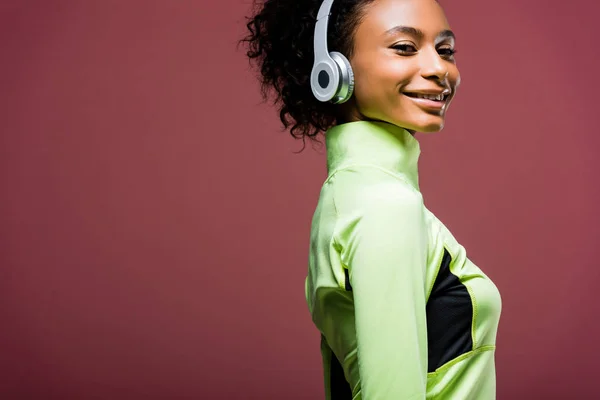 Красива усміхнена афроамериканська спортсменка в навушниках, дивлячись на камеру ізольована на коричневому з місцем для тексту — стокове фото