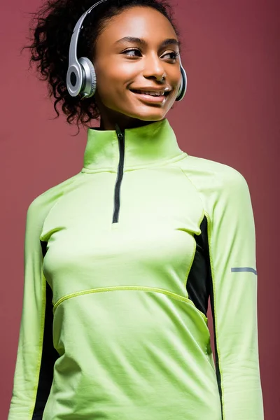 Красивая улыбающаяся африканская спортсменка в наушниках и беговой куртке, изолированная на коричневом — стоковое фото