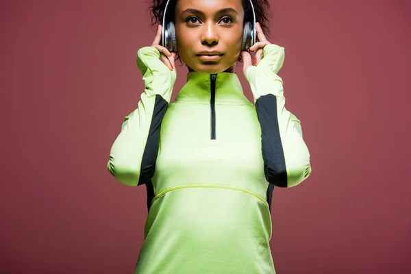 Bela esportista afro-americana em fones de ouvido e jaqueta de pista isolada em marrom — Fotografia de Stock