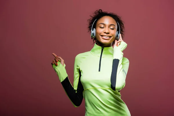 Bela sorridente afro-americana esportista em fones de ouvido ouvir música com os olhos fechados isolado em marrom — Fotografia de Stock