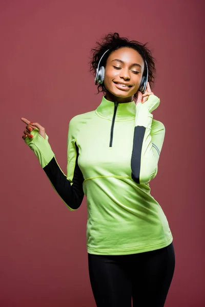 Beautiful african american sportswoman in headphones listening music with eyes closed isolated on brown — Stock Photo