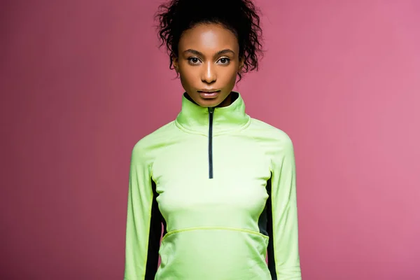 Hermosa afroamericana deportista en chaqueta de atletismo mirando a la cámara aislada en rosa - foto de stock