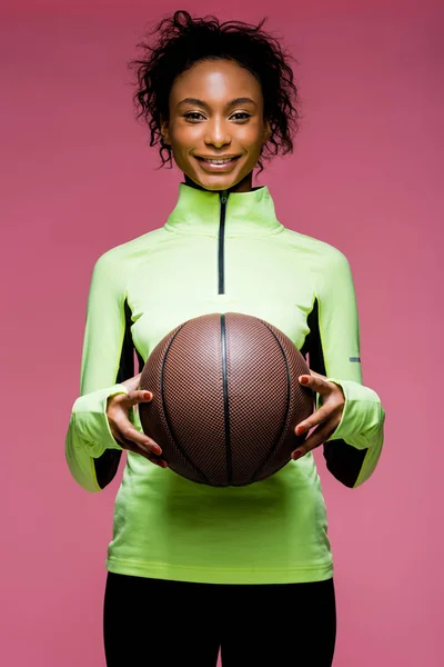 Schöne lächelnde afrikanisch-amerikanische Sportlerin blickt in die Kamera und hält Basketball isoliert auf rosa — Stockfoto