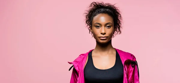 Prise de vue panoramique d'une jolie sportive afro-américaine en coupe-vent regardant une caméra isolée sur rose avec espace de copie — Photo de stock