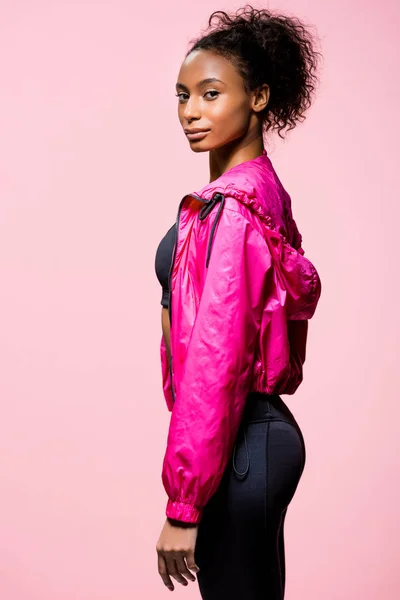 Attractive african american sportswoman in windbreaker looking at camera and posing isolated on pink — Stock Photo