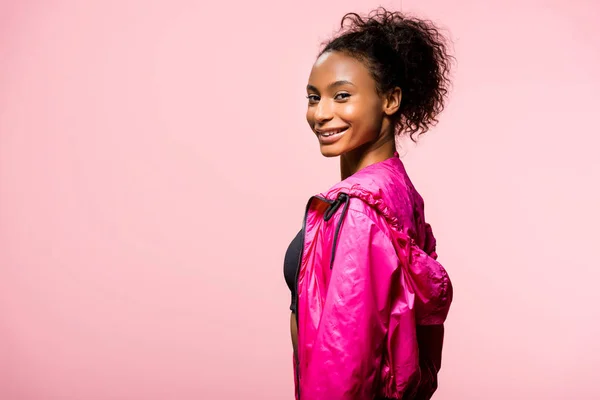 Attractive african american sportswoman in windbreaker looking at camera and smiling isolated on pink with copy space — Stock Photo