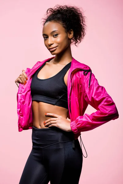 Attraente sportiva africana americana in giacca a vento con mano sul fianco guardando la fotocamera isolata sul rosa — Foto stock