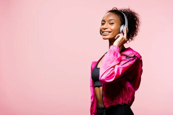 Красива усміхнена афроамериканська спортсменка в навушниках, дивлячись на камеру ізольована на рожевому з копіювальним простором — стокове фото