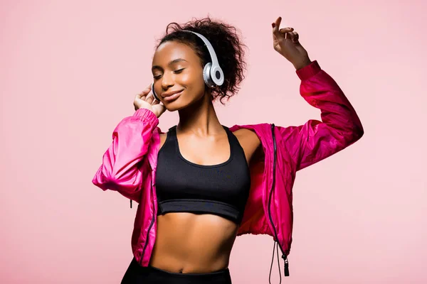 Schöne afrikanisch-amerikanische Sportlerin in Kopfhörer Musik hören und gestikulieren isoliert auf rosa — Stockfoto