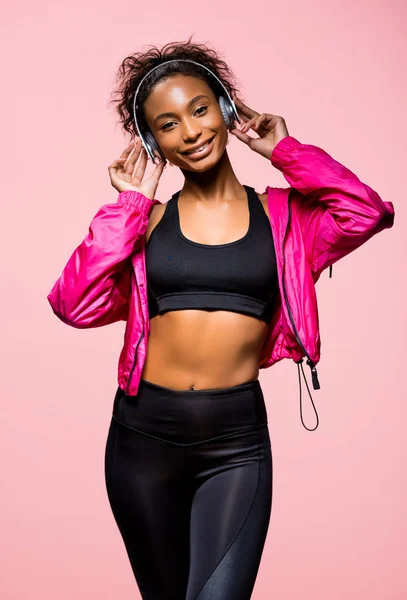 Hermosa afroamericana deportista en auriculares escuchando música y mirando a la cámara aislada en rosa - foto de stock