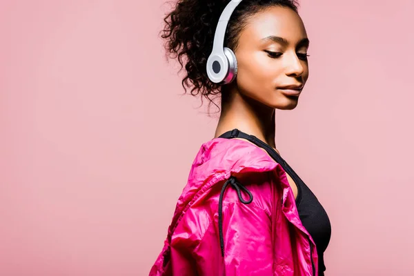 Hermosa deportista afroamericana en cortavientos y auriculares aislados en rosa con espacio de copia - foto de stock