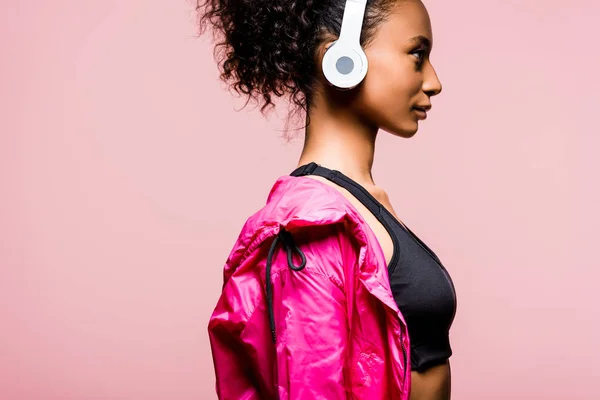 Vue latérale de la belle sportive afro-américaine en coupe-vent et écouteurs isolés sur rose — Photo de stock