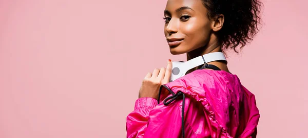 Plan panoramique de belle sportive afro-américaine avec écouteurs regardant la caméra isolée sur rose — Photo de stock