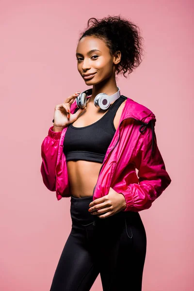 Hermosa afroamericana deportista con auriculares mirando a la cámara y sonriendo aislado en rosa - foto de stock