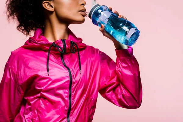 Обрезанный вид на африканскую американскую спортсменку, пьющую воду из спортивной бутылки, изолированной на розовый — стоковое фото