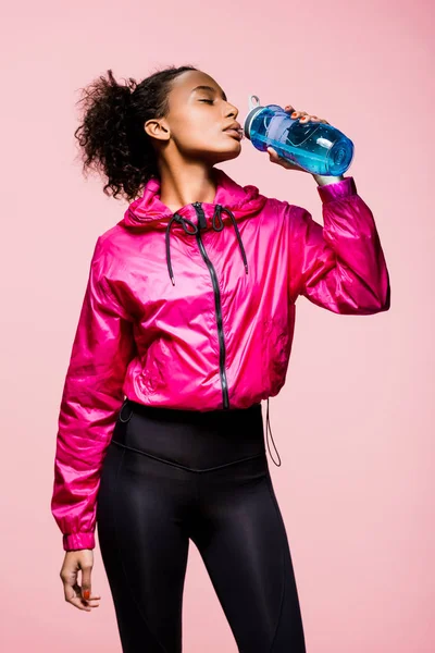 Beautiful african american sportswoman drinking water from sport bottle isolated on pink — Stock Photo