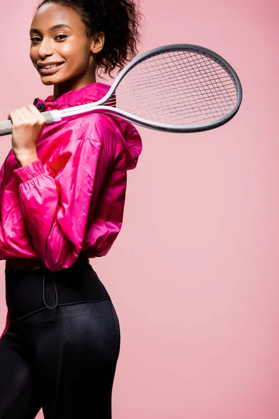 Bella sportiva afroamericana guardando la macchina fotografica mentre posa con racchetta da tennis isolata su rosa — Foto stock