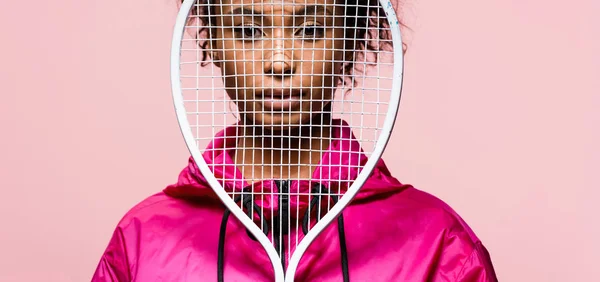 Plano panorámico de hermosa deportista afroamericana cubriendo la cara con raqueta de tenis y mirando a la cámara aislada en rosa - foto de stock
