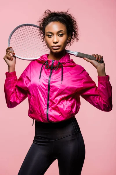 Hermosa deportista afroamericana con raqueta de tenis posando y mirando a la cámara aislada en rosa - foto de stock