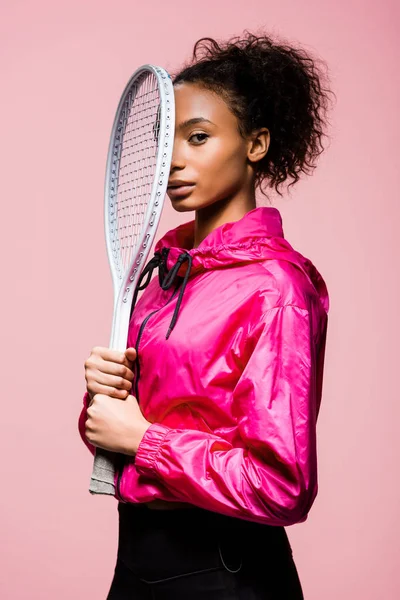 Bella sportiva afro-americana in possesso di racchetta da tennis e guardando la fotocamera isolata su rosa — Foto stock