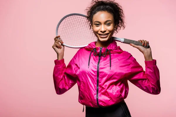 Bellissima sportiva afroamericana che tiene in mano la racchetta da tennis, guarda la macchina fotografica e sorride isolata sul rosa — Foto stock