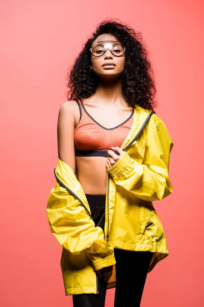 Bela esportista afro-americana em óculos olhando para a câmera e posando isolado em coral — Fotografia de Stock