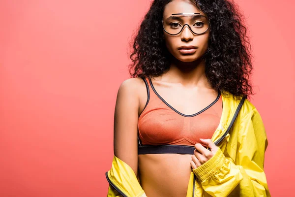 Hermosa deportista afroamericana en gafas mirando a la cámara y posando aislado en coral con espacio de copia - foto de stock