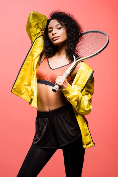 Belle sportive afro-américaine regardant la caméra et tenant une raquette de tennis isolée sur du corail — Photo de stock