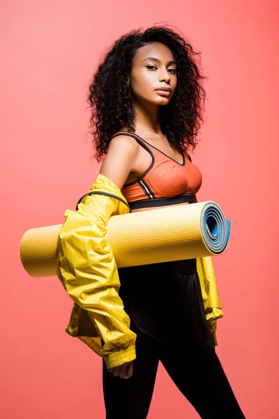 Beautiful african american sportswoman looking at camera and holding sport mat isolated on coral — Stock Photo