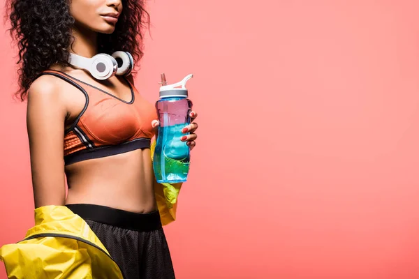 Обрізаний вид афроамериканської спортсменки з навушниками та спортивною пляшкою ізольовано на коралі з місцем для тексту — стокове фото
