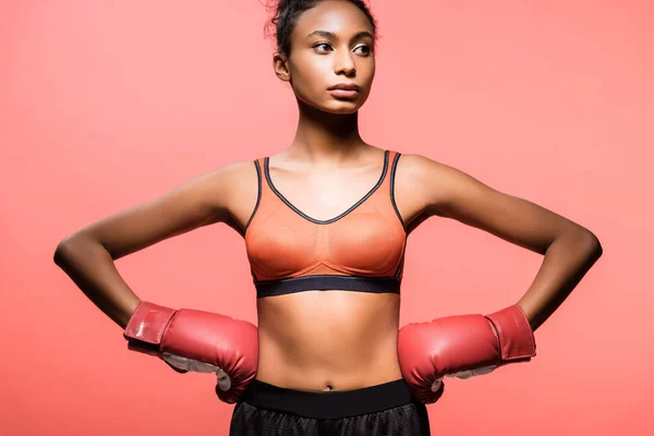 Belle sportive afro-américaine en gants de boxe posant avec les mains akimbo isolé sur du corail — Photo de stock