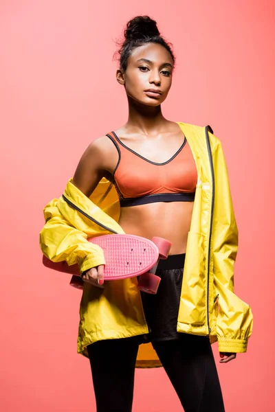 Beautiful african american sportswoman holding penny board and looking at camera isolated on coral — Stock Photo