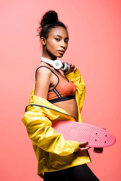 Beautiful african american sportswoman with headphones looking at camera while holding penny board isolated on coral — Stock Photo